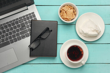 Laptop with notepad and cup of tea with cookies on blue wooden background