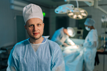 Closeup portrait of male doctor in surgical uniforms standing posing looking at camera in dark modern operating room. Diverse team of surgeons performing operation on background