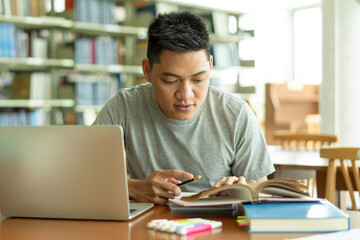 male asian student studying and reading book in library - obrazy, fototapety, plakaty