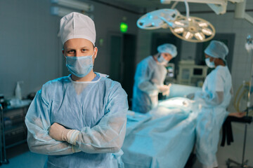 Portrait of male doctor in surgical uniforms and masks standing posing looking at camera with folded arms in operating theater. Diverse professional team of surgeons performing operation on background