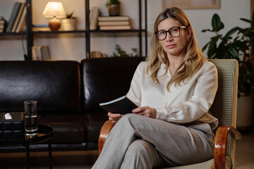 Female psychotherapist sitting in armchair at her workplace holding notebook and looking at camera