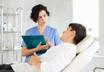 Middle-aged female doctor signing papers while talking with man patient lying on clinical chair