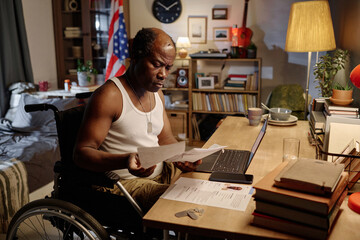 Serious african american man in wheelchair sitting at desk in bedroom and looking through documents