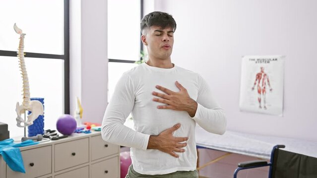 At his balance point, a portrait of a young, handsome hispanic man finding calm by meditating and taking a deep breath, relaxing in the indoor room of a rehab clinic, conquering respiratory problems