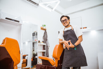 asian male hairstylist leaning on barber chair and looking at camera with smiling face in hair salon
