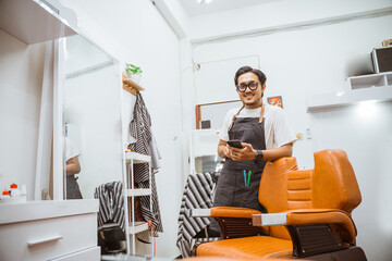 asian male hairdresser looking at camera with happy expression while holding phone and standing beside barber chair in hair salon