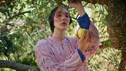 Sensual fashion model standing under green tree branches holding apple close up.