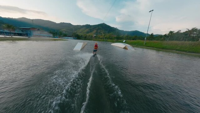 Beginner wakeboarder man in orange construction helmet trying cable wakeboard in lake park water sport. Extreme activity guy training practicing wakeboarding water sport fpv drone follow aerial shot