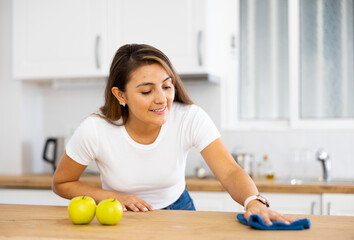 Smiling young Hispanic woman wiping dust from kitchen surfaces with cleaning rag and detergent spray