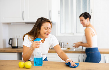 Fototapeta premium Positive young Latina wiping table with cloth and spray while cleaning kitchen with sister. Everyday household chores concept