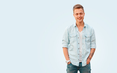 Happy handsome young guy with a hairstyle in a denim fashion shirt and a white T-shirt with jeans stands on an isolated blue background