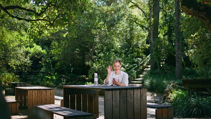 Fototapeta premium Beautiful woman greeting smartphone camera sitting wooden table in green park. 