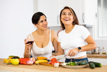 Obraz na płótnie Canvas Two cheerful young Hispanic female roommates enjoying weekend together, drinking wine and preparing vegetable dish for dinner at home kitchen