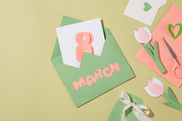 An envelope with a card printed the number 8 inside and the word “March” decorated outside. A scissor arranged with some cards, paper tulip flowers and paper flowers