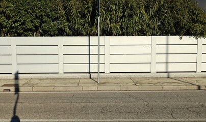 Pole on concrete sidewalk with fence made of white horizontal aluminium slats and hedge on behind....