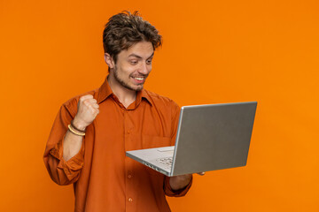 Happy excited Caucasian man typing on laptop, working on project, enjoying results, winning lottery game celebrating success, online shopping victory good email news. Handsome guy on orange background