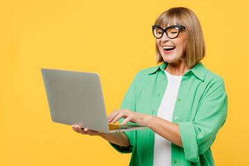 Elderly smiling happy blonde IT woman 50s years old wear green shirt glasses casual clothes hold use work on laptop pc computer isolated on plain yellow background studio portrait. Lifestyle concept.