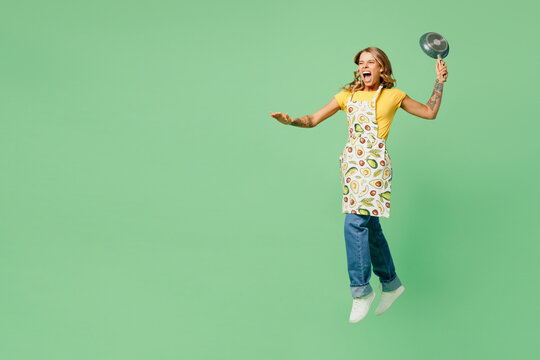 Full body young angry mad furious outraged housewife housekeeper chef cook baker woman wearing apron yellow t-shirt jump high with frying pan isolated on plain green background. Cooking food concept.