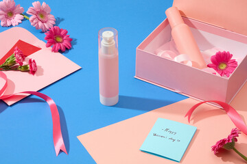 Close up of a set of pink cosmetics, a card with the words happy Women's day, gerberas, carnations and ribbons are displayed on a blue background.