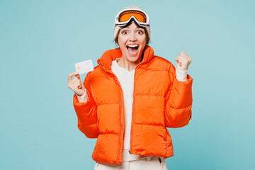 Skier young woman wears padded windbreaker jacket hat ski goggles mask hold credit bank card do winner gesture travel rest spend weekend winter season in mountains isolated on plain blue background. © ViDi Studio