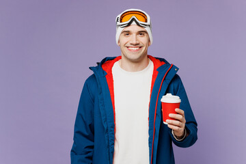 Skier fun man wear warm blue windbreaker jacket ski goggles mask hat hold takeaway delivery paper cup coffee to go spend extreme weekend winter season in mountains isolated on plain purple background.