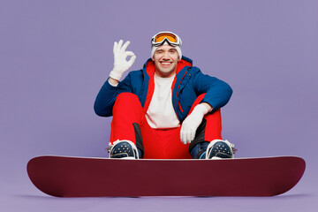 Full body happy man wear blue windbreaker jacket ski goggles mask hat sitting with snowboard show...