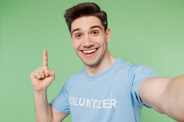 Young smart man wear blue t-shirt white title volunteer doing selfie shot pov mobile cell phone point finger up isolated on plain pastel green background. Voluntary free team work help grace concept.
