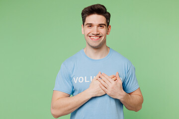 Young grateful happy man wears blue t-shirt white title volunteer put folded hands on heart isolated on plain pastel light green background. Voluntary free work assistance help charity grace concept.