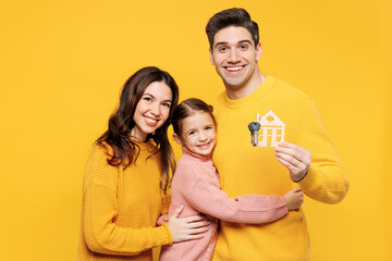 Young happy fun parents mom dad with child kid girl 7-8 years old wear pink sweater casual clothes hold keys and wooden house mockup hug cuddle isolated on plain yellow background. Family day concept.