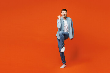 Fototapeta na wymiar Full body smiling happy overjoyed young man he wears blue shirt white t-shirt casual clothes do winner gesture raise up leg isolated on plain red orange background studio portrait. Lifestyle concept.