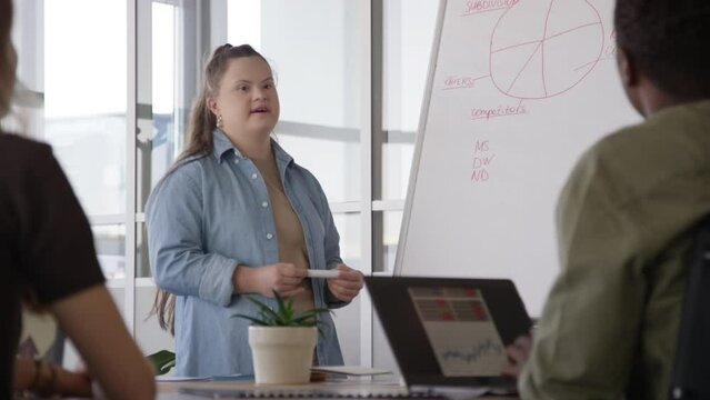 Medium shot of young Caucasian female trainer with down syndrome standing beside flipchart and answering questions of co-workers during seminar meeting in office conference room