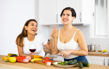 Obraz na płótnie Canvas Smiling young Hispanic woman cooking with sister in home kitchen, chatting cheerfully and drinking wine while preparing vegetable salad. Happy family relationship concept..