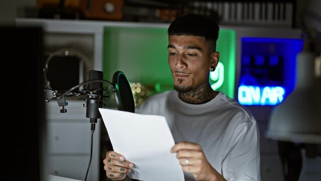 Serious young latin man, tattooed musician reading his song from a music sheet, singing his heart out in a dimly lit music studio