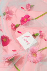 Top view of a pastel blue mesh fabric overlaid on a pink background. Carnations and gerberas are decorated around a card with the word Love.