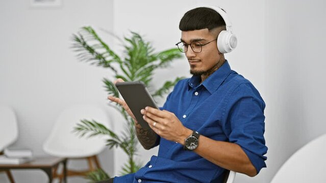 Focused young latin man immersed in work on his touchpad amidst calming song rhythms in the waiting room, his serious gaze accentuated by his glasses