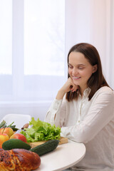 Obraz na płótnie Canvas Caucasian brown haired woman sitting at kitchen table with fruit and vegetables having ingredients delightful and nutritious dish holding chin enjoying cooking and healthy nutrition
