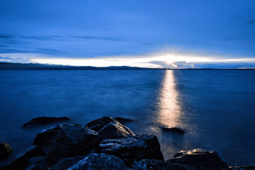 Sonnenuntergang am Bodensee. Sonnenstrahl reflektiert im Wasser zu einem Lichtstrahl. Große dunkle Steine im Vordergrund. Dunkelblaues Wasser im See. Blauber bedeckter Himmel - obrazy, fototapety, plakaty