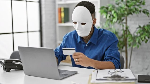 Masked young latin man pulls off a daring credit card hack on his laptop at the office, exposing the dark underbelly of internet crime