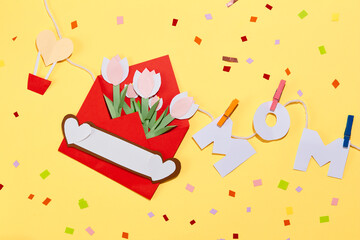 Paper tulips inside a red envelope, the word MOM hung on a string with wooden clips and colorful confetti on a yellow background. Women's day theme for advertising.