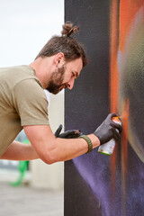 Attractive male artist is painting picture with paint spray can spraying it onto canvas at outdoor street exhibition, side view of man art maker