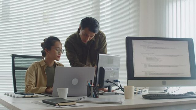 Medium long shot of two Asian coders analyzing idea while developing new app for users using computer during work in office