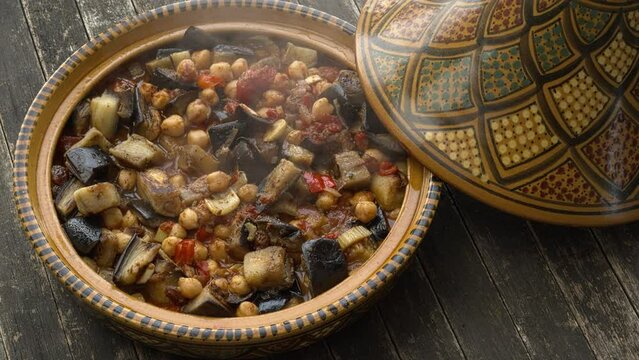 Traditional Moroccan tagine pot standing on a rustic wooden table that has just been opened. Boiling vegan tagine made of eggplant and chickpea inside. Zooming in.
