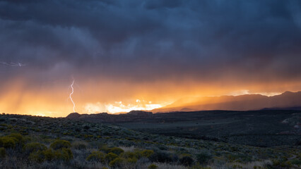Lightning strikes from the dark clouds above Pine Valley Mt. through the brilliant orange clouds lit by the setting sun to the shadowed landscape of Southern Utah during a late season monsoon storm.