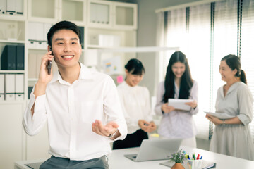 Young Asian business man using smartphone while sitting on table in office with smile. Man in white shirt looking away while talking to someone on mobilephone with co-worker background.