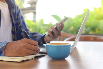 Close up of male hand writing on note book with pen, showing blank screen of laptop and smartphone. Working in cafe concept with a cup of coffee