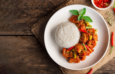 Top view of rice topped with stir fried crispy pork with red curry in white plate on wooden table background. Thai Food