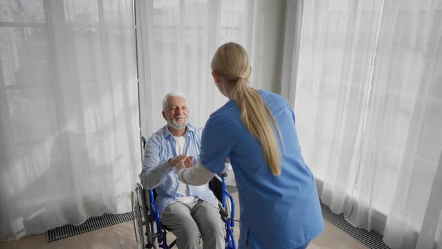 Nurse talking senior man in wheelchair. Female social welfare physical therapist looks after a senior male, providing medical care at clinic. Living with legs paralysis, special needs people.