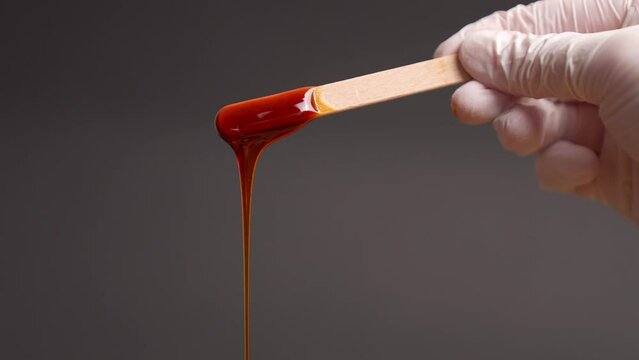 Pouring liquid brown sugar paste or wax for depilation on a stick close-up hands in white latex gloves