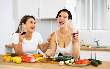 Obraz na płótnie Canvas Happy female friends with glasses of red wine chatting and preparing salad together in modern kitchen