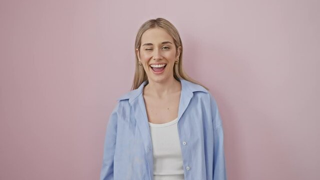 Flirty fun as young blonde woman winks at the camera with a sexy expression! blonde bombshell wearing a shirt looking cheerful with a happy face, over an isolated pink background.